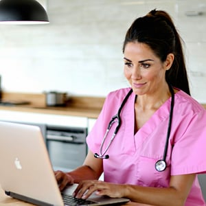 a nurse applying for a job wearing pink scrubs sat at a laptop scrolling job vacancies Sat a kitchen table looking at the screen-1